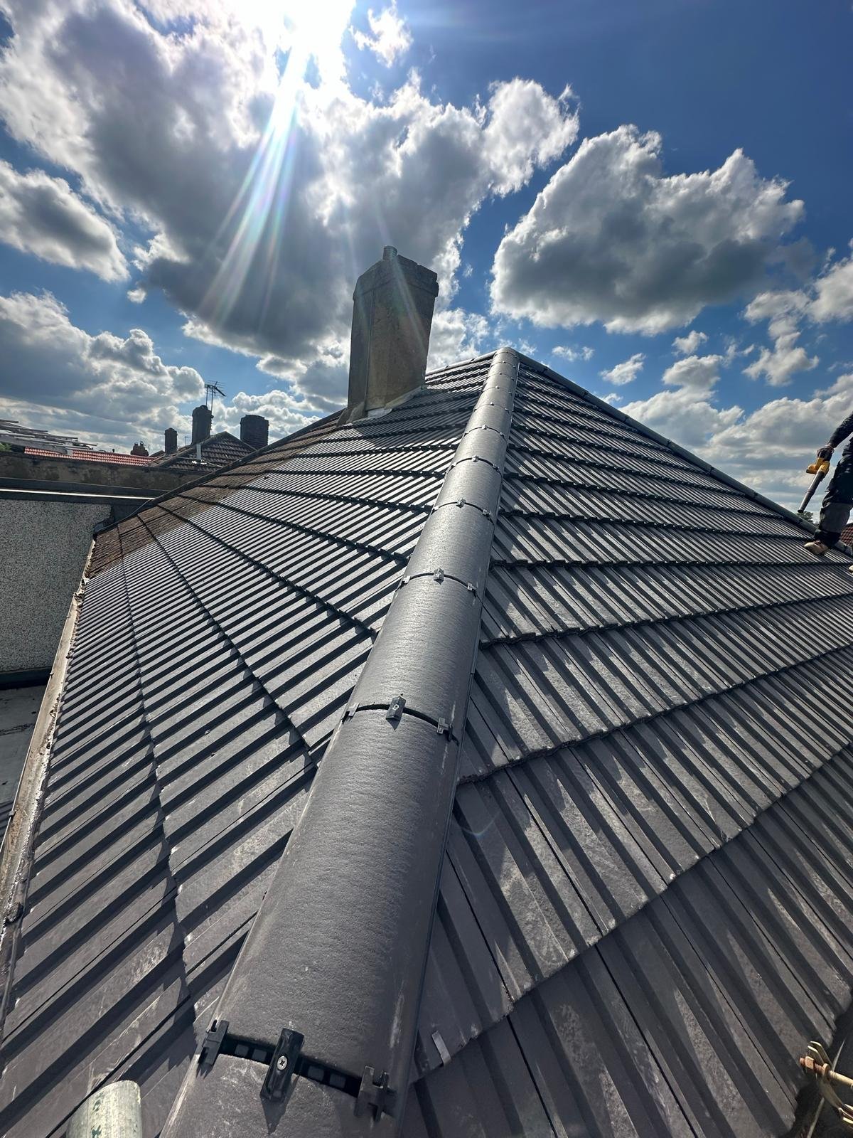 Grey metal-effect corrugated tile roof with a ridge line and sun breaking through cloud behind