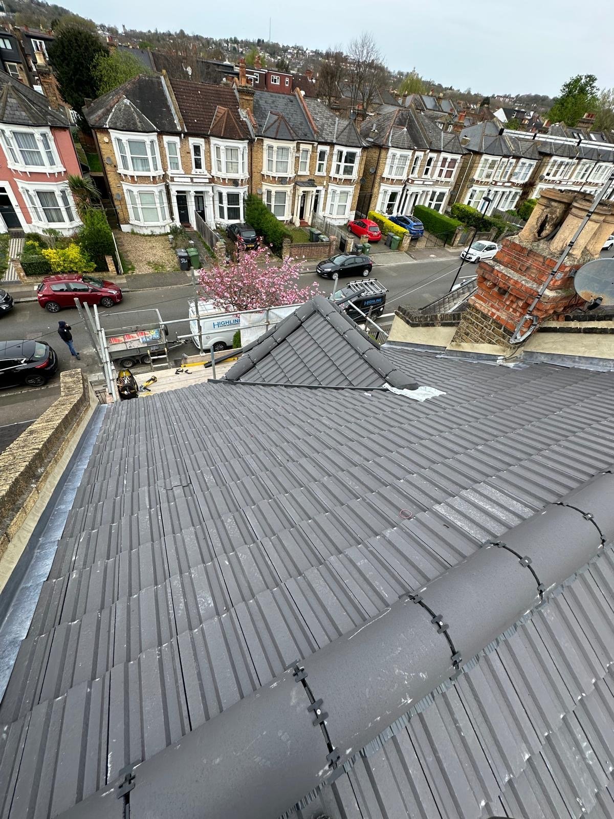 Completed dark grey tile re-roof over a row of Victorian terraces, viewed from scaffolding with parked cars below